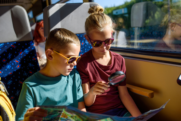 Siblings sitting in train, looking at map with magnifying glass. Family going on hike, using a public transport.