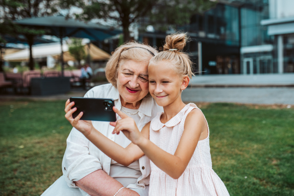 Grandma taking selfie with granddaugter sitting on bench. Girl spending time with senior grandmother, during summer break or after school.