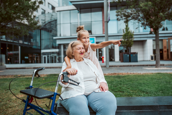 Grandma with walker and granddaugter in the city, sitting on bench. Girl spending time with senior grandmother, during holidays, after school.