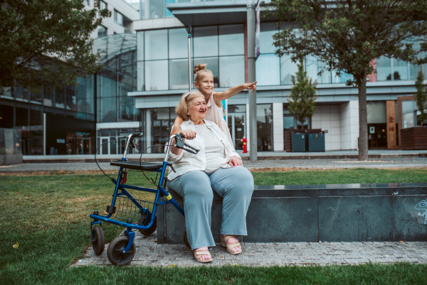 Grandma with walker and granddaugter in the city, sitting on bench. Girl spending time with senior grandmother, during holidays, after school.