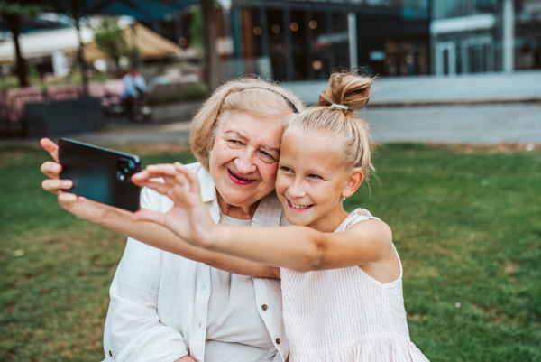 Grandma taking selfie with granddaugter sitting on bench. Girl spending time with senior grandmother, during summer break or after school.