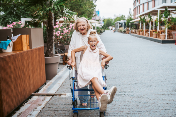 Girl sitting on rollator, grandma pushing her, laughing. Granddaughter spending time with senior grandmother, during summer break or after school.