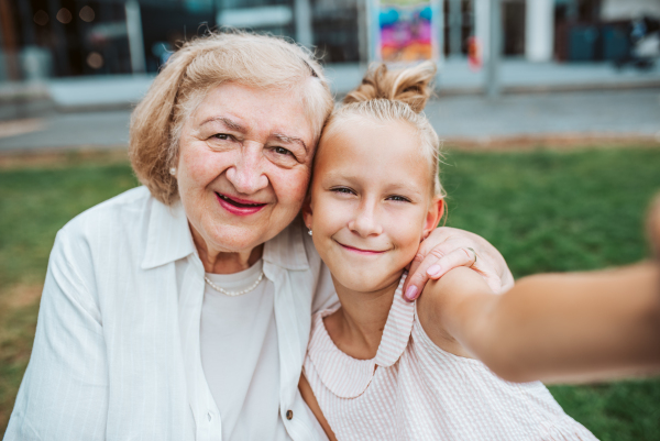 Grandma taking selfie with granddaugter sitting on bench. Girl spending time with senior grandmother, during summer break or after school.