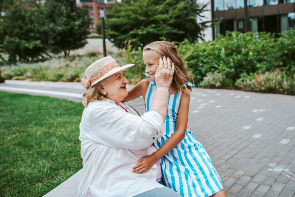 Grandma sitting on bench with granddaugter in the city, caressing her face. Girl spending time with senior grandmother, during summer break or after school.