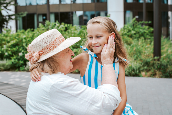 Grandma sitting on bench with granddaugter in the city, caressing her face. Girl spending time with senior grandmother, during summer break or after school.