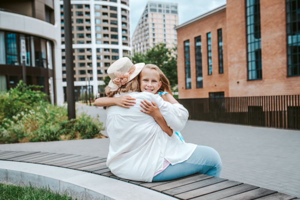 Grandma sitting on bench with granddaugter in the city, embracing her. Girl spending time with senior grandmother, during summer break or after school.