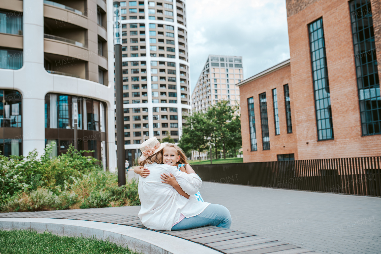 Grandma sitting on bench with granddaugter in the city, embracing her. Girl spending time with senior grandmother, during summer break or after school.