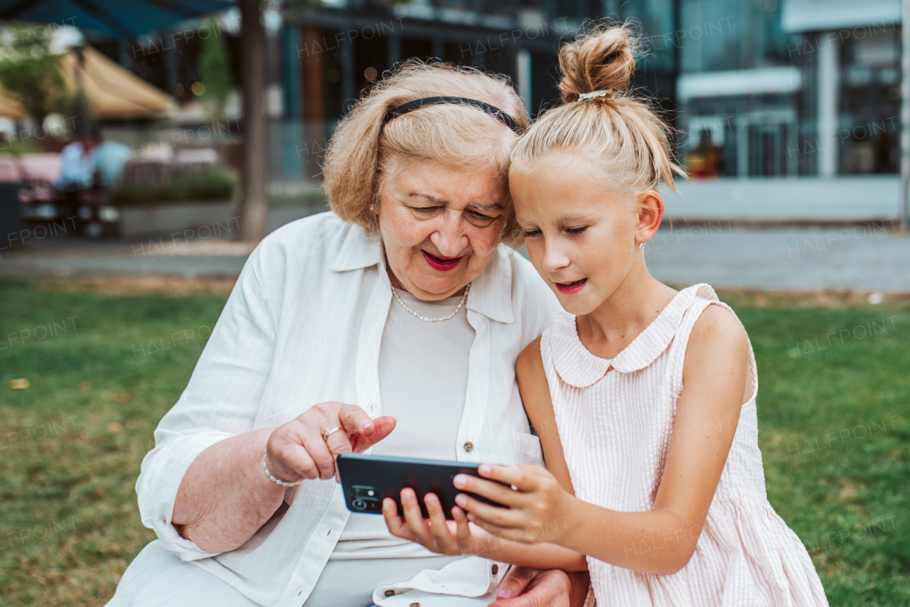 Grandma picking up young girl from school at afternoon. Granddaughter showing something on smartphone to senior grandmother.