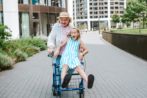 Grandma pushing girl sitting on rollator, having fun. Granddaughter spending time with senior grandmother, during summer break or after school.