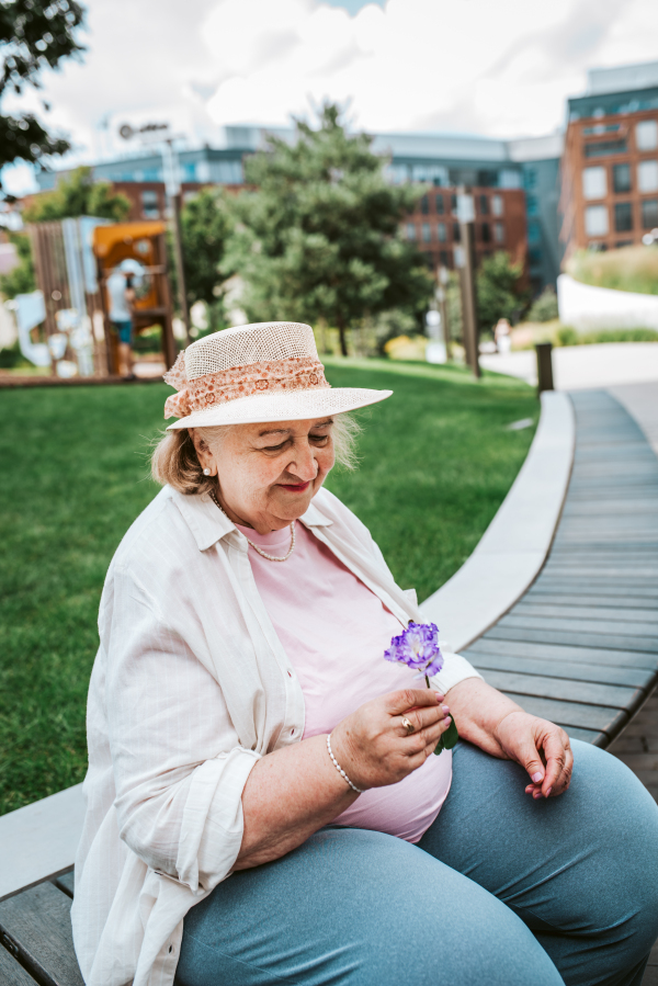 Elderly woman sitting on bench, holding single purple flower. Senior woman running errands, feeling fatigued.