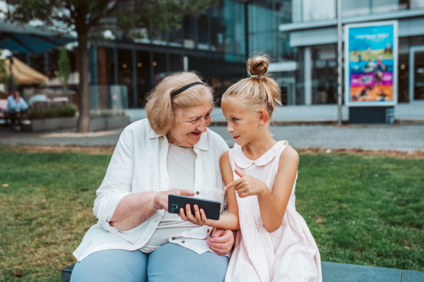 Grandma picking up young girl from school at afternoon. Granddaughter showing something on smartphone to senior grandmother.