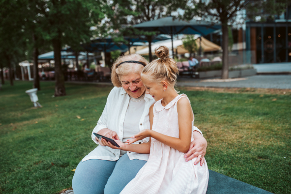 Grandma picking up young girl from school at afternoon. Granddaughter showing something on smartphone to senior grandmother.