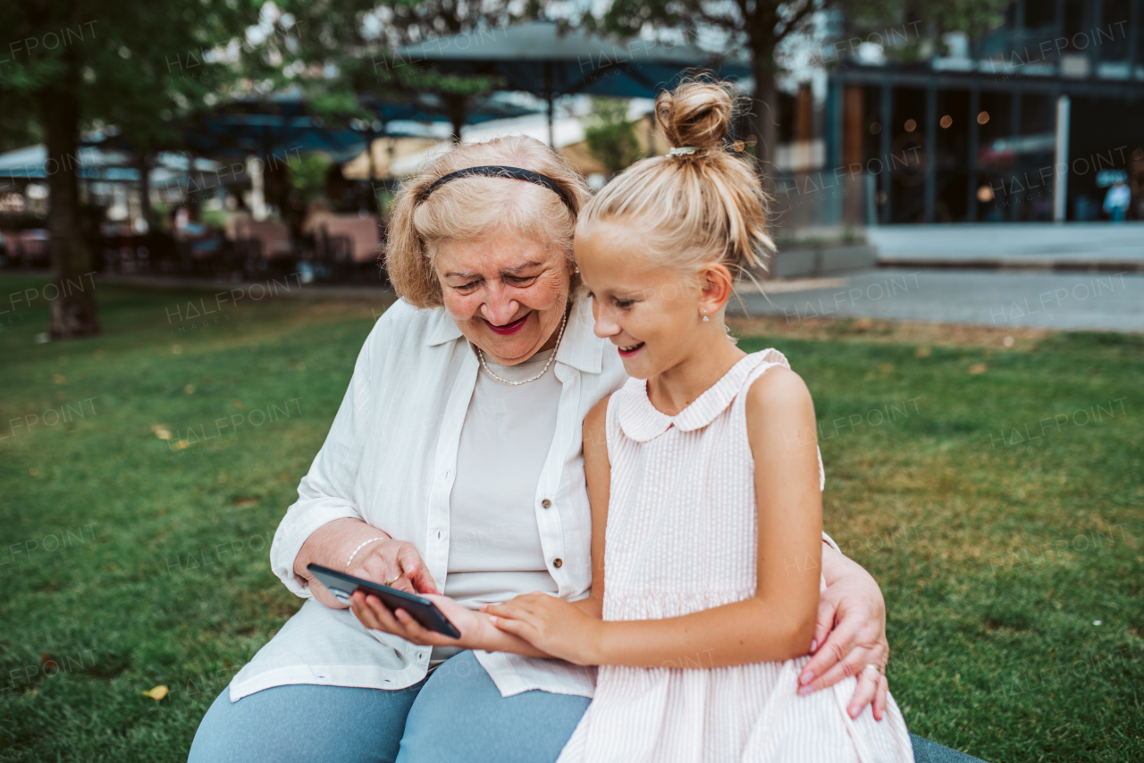 Grandma picking up young girl from school at afternoon. Granddaughter showing something on smartphone to senior grandmother.