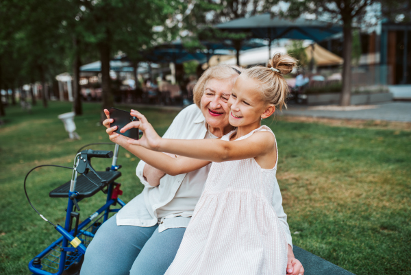 Grandma taking selfie with granddaugter sitting on bench. Girl spending time with senior grandmother, during summer break or after school.