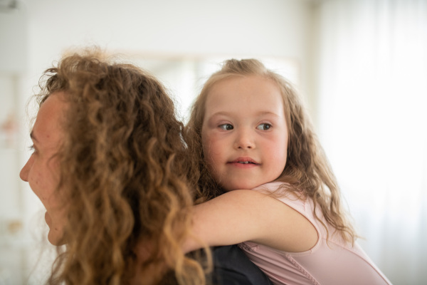 Young happy mother holding her daughter with down syndome piggyback, having fun in a ballet dance studio, concept of education and integration dissabled children.