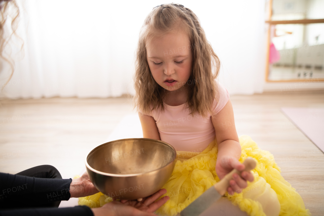 Little girl with down syndrom sitting on the floor in a ballet dancing studio and making sound on tibetian singing bowl.