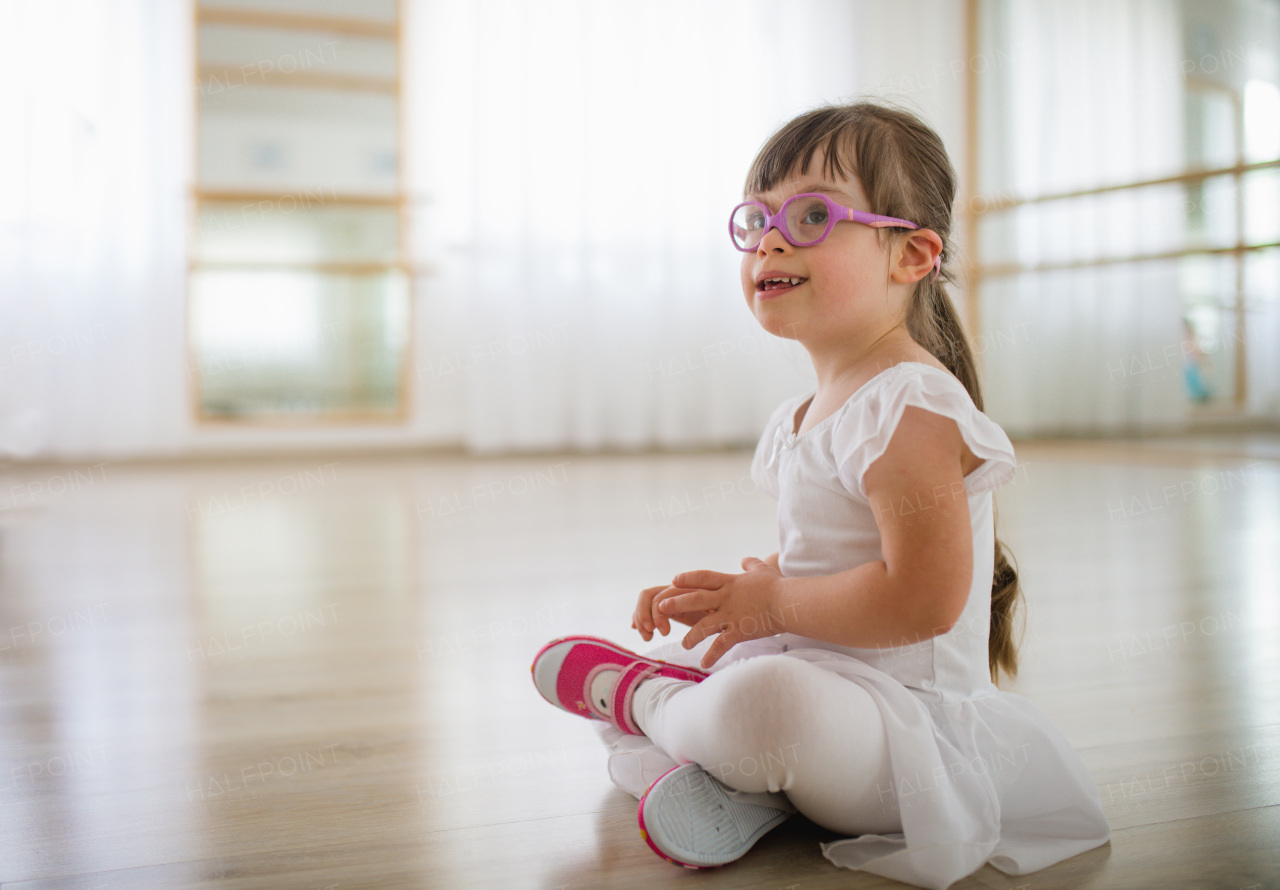 Little girl with down syndrome at a ballet class in dance studio, sitting and resting. Concept of integration and education of disabled children.