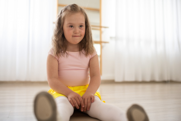 Little girl with down syndrome at a ballet class in dance studio, sitting and resting. Concept of integration and education of disabled children.