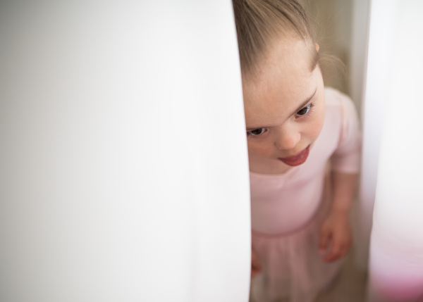 High angle view of little girl with down syndrome hidding behind a curtain, at ballet class in dance studio. Concept of integration and education of disabled children.