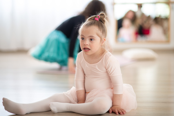 Little girl with down syndrome at a ballet class in dance studio, sitting and resting. Concept of integration and education of disabled children.