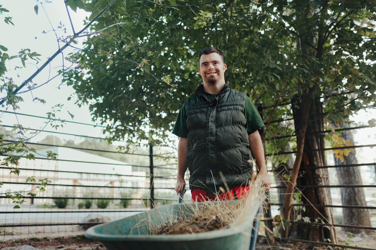 Caretaker with down syndrome in the zoo pushing wheelbarrow after tidying animal enclosure. Concept of integration people with disabilities into society.