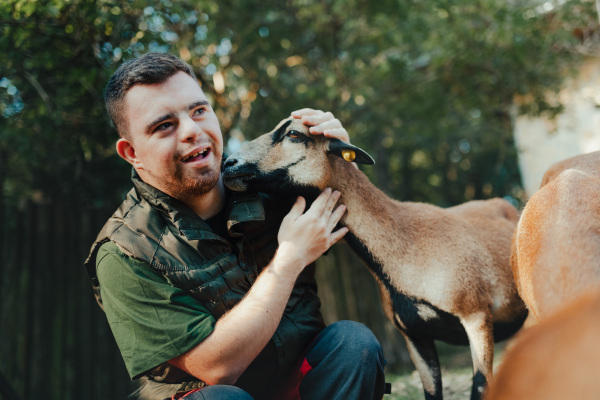 Caretaker with down syndrome taking care of animals in zoo, stroking a goat. Concept of integration people with disabilities into society.