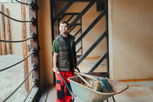 Caretaker with down syndrome in the zoo pushing wheelbarrow after tidying animal enclosure. Concept of integration people with disabilities into society.