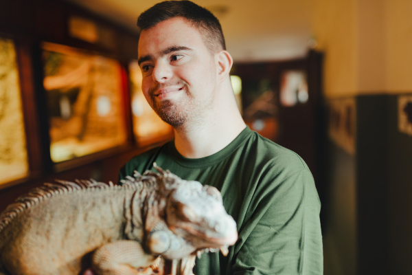 Portrait of caretaker with down syndrome taking care of animals in zoo, stroking an iguana. Concept of integration people with disabilities into society.