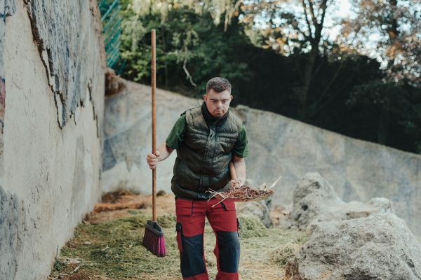 Caretaker with down syndrome in the cleaning animal enclosure. Concept of integration people with disabilities into society.