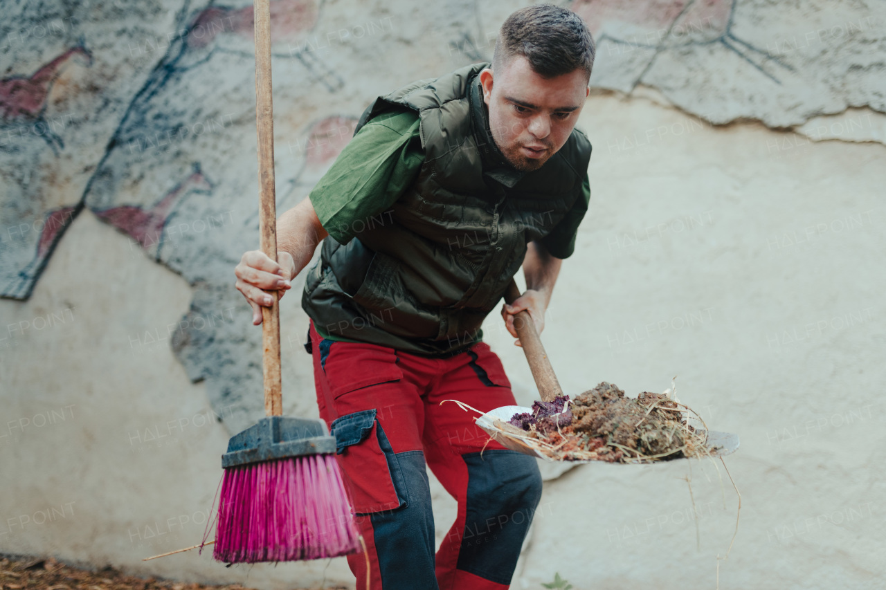 Caretaker with down syndrome in the cleaning animal enclosure. Concept of integration people with disabilities into society.