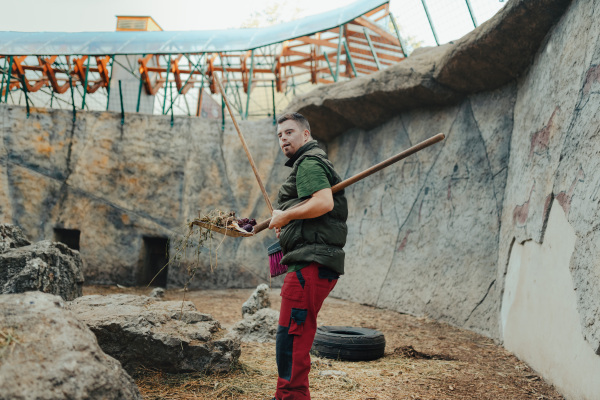 Caretaker with down syndrome in the cleaning animal enclosure. Concept of integration people with disabilities into society.