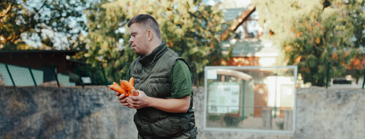 Caretaker with down syndrome in the giving carrot in animal enclosure. Concept of integration people with disabilities into society.