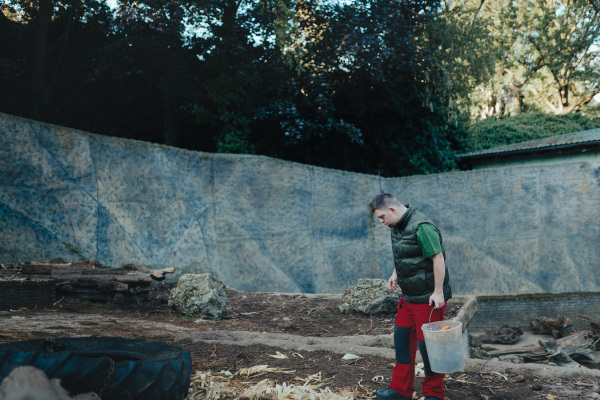 Caretaker with down syndrome in the zoo giving food in animal enclosure. Concept of integration people with disabilities into society.