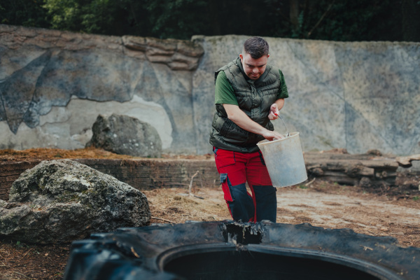 Caretaker with down syndrome in the zoo giving food in animal enclosure. Concept of integration people with disabilities into society.