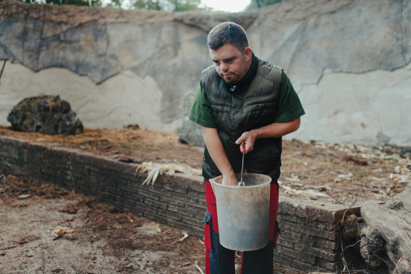 Caretaker with down syndrome in the zoo giving food in animal enclosure. Concept of integration people with disabilities into society.