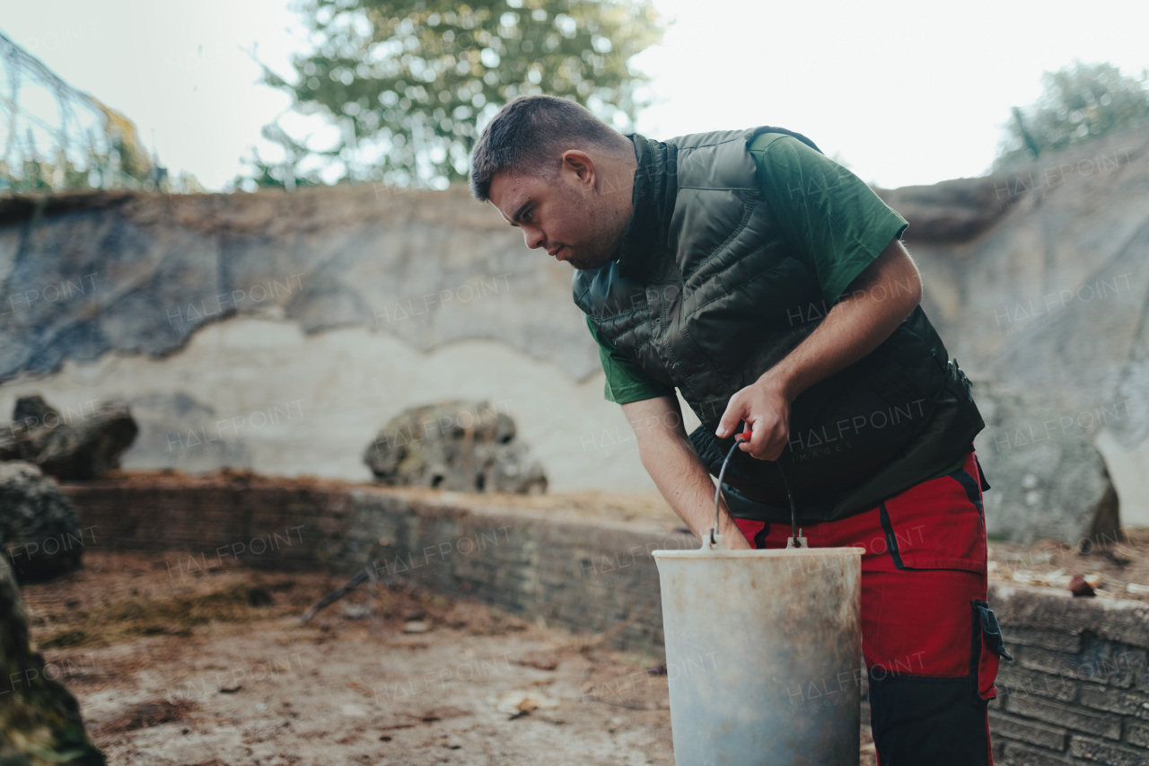 Caretaker with down syndrome in the zoo giving food in animal enclosure. Concept of integration people with disabilities into society.