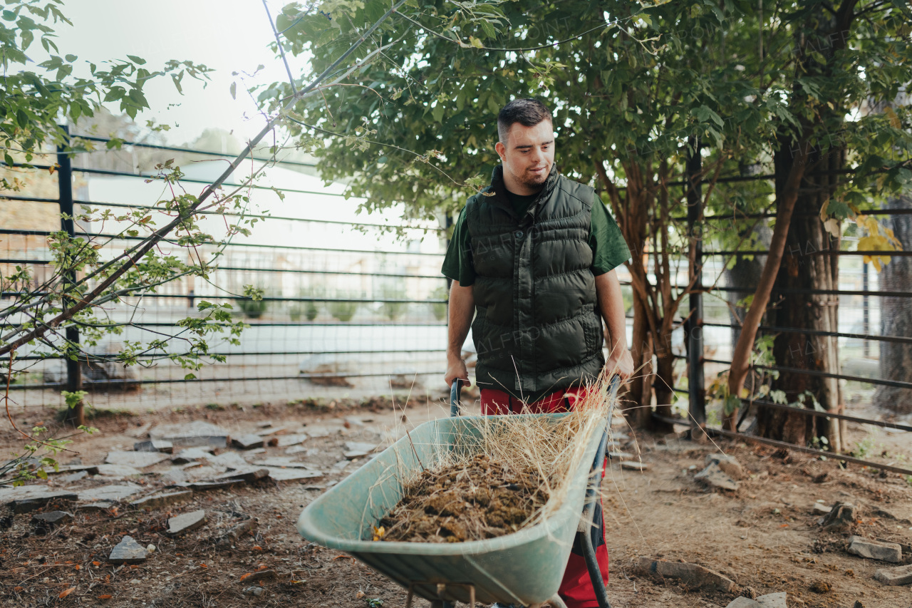 Caretaker with down syndrome in the zoo pushing wheelbarrow after cleaning animal enclosure. Concept of integration people with disabilities into society.