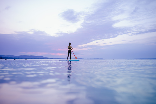 A young beautiful girl surfer paddling on surfboard on the lake at sunrise