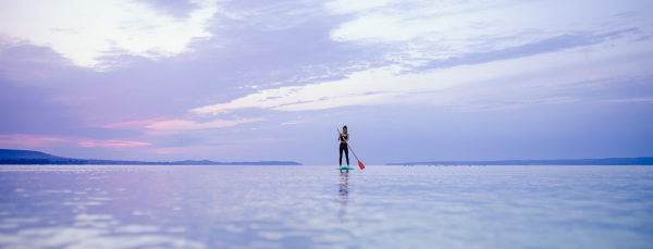 A young beautiful girl surfer paddling on surfboard on the lake at sunrise