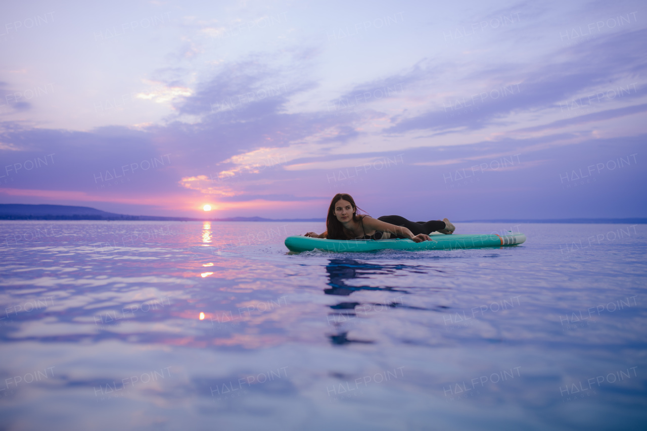 A young beautiful girl surfer paddling on surfboard on the lake at sunrise