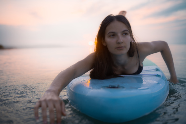 A young beautiful girl surfer paddling on surfboard on the lake at sunrise