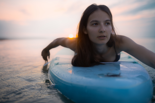 A young beautiful girl surfer paddling on surfboard on the lake at sunrise