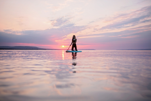 A young beautiful girl surfer paddling on surfboard on the lake at sunrise