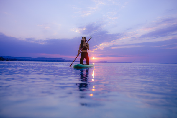 A young beautiful girl surfer paddling on surfboard on the lake at sunrise