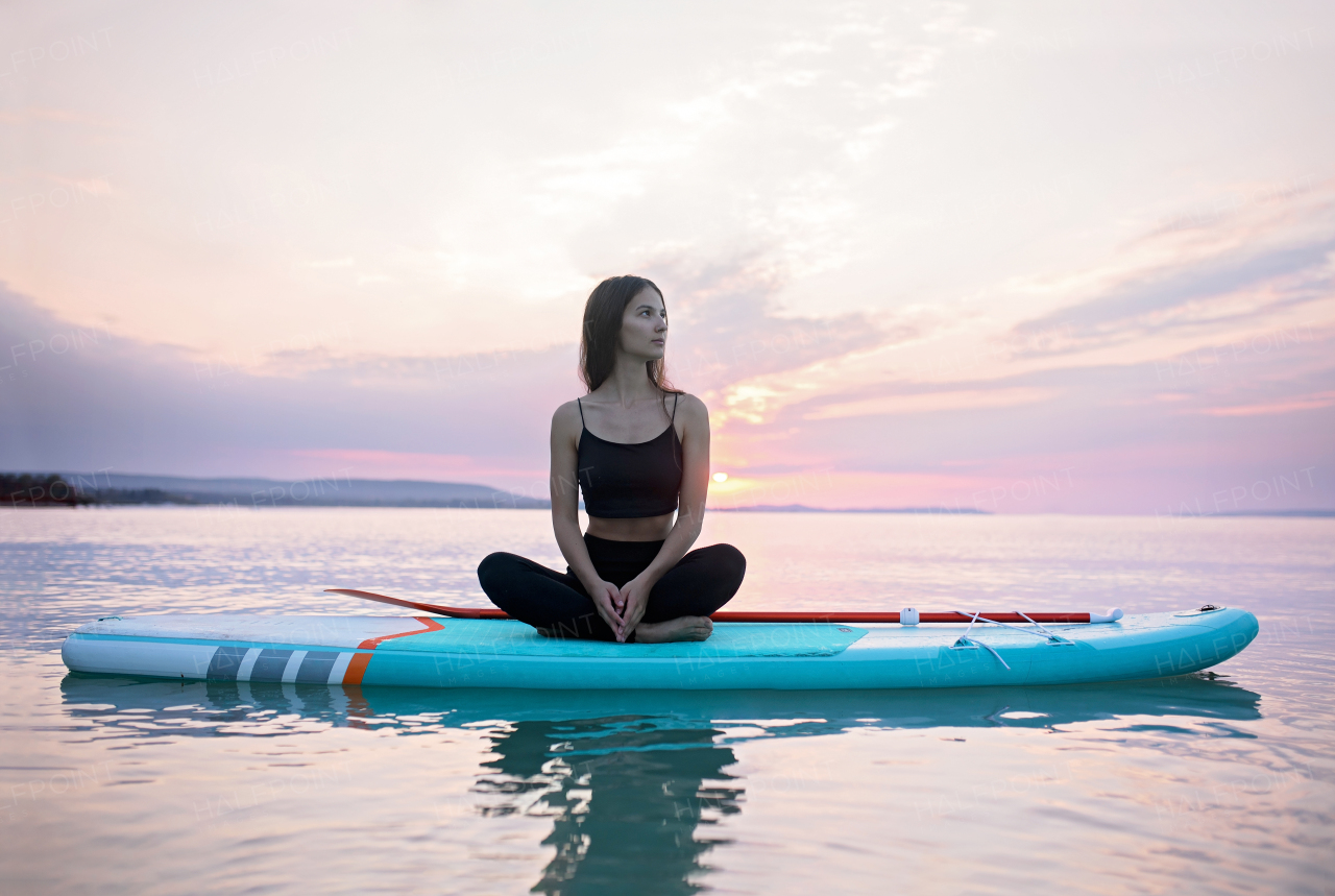 A young surfer girl meditating on surfboard on the lake at sunrise.