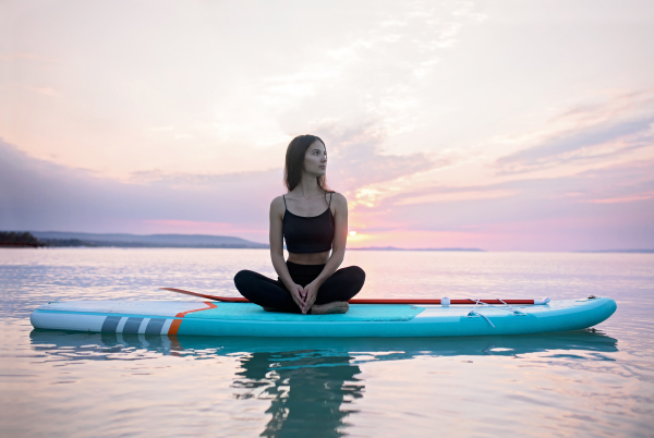 A young surfer girl meditating on surfboard on the lake at sunrise.