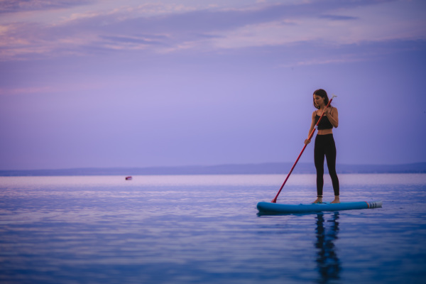 A young beautiful girl surfer paddling on surfboard on the lake at sunrise