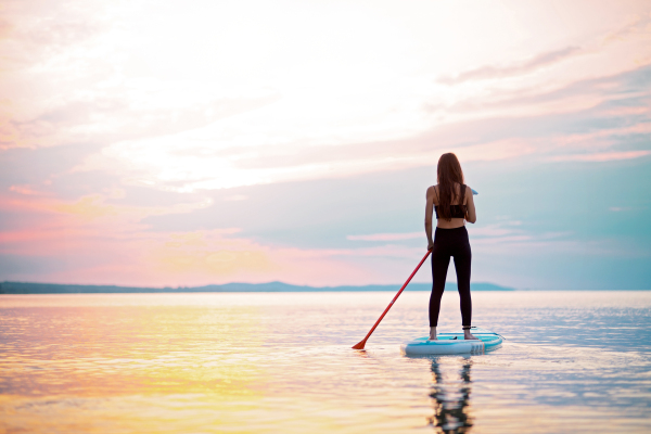 A rear view of girl surfer paddling on surfboard on the lake at sunrise.