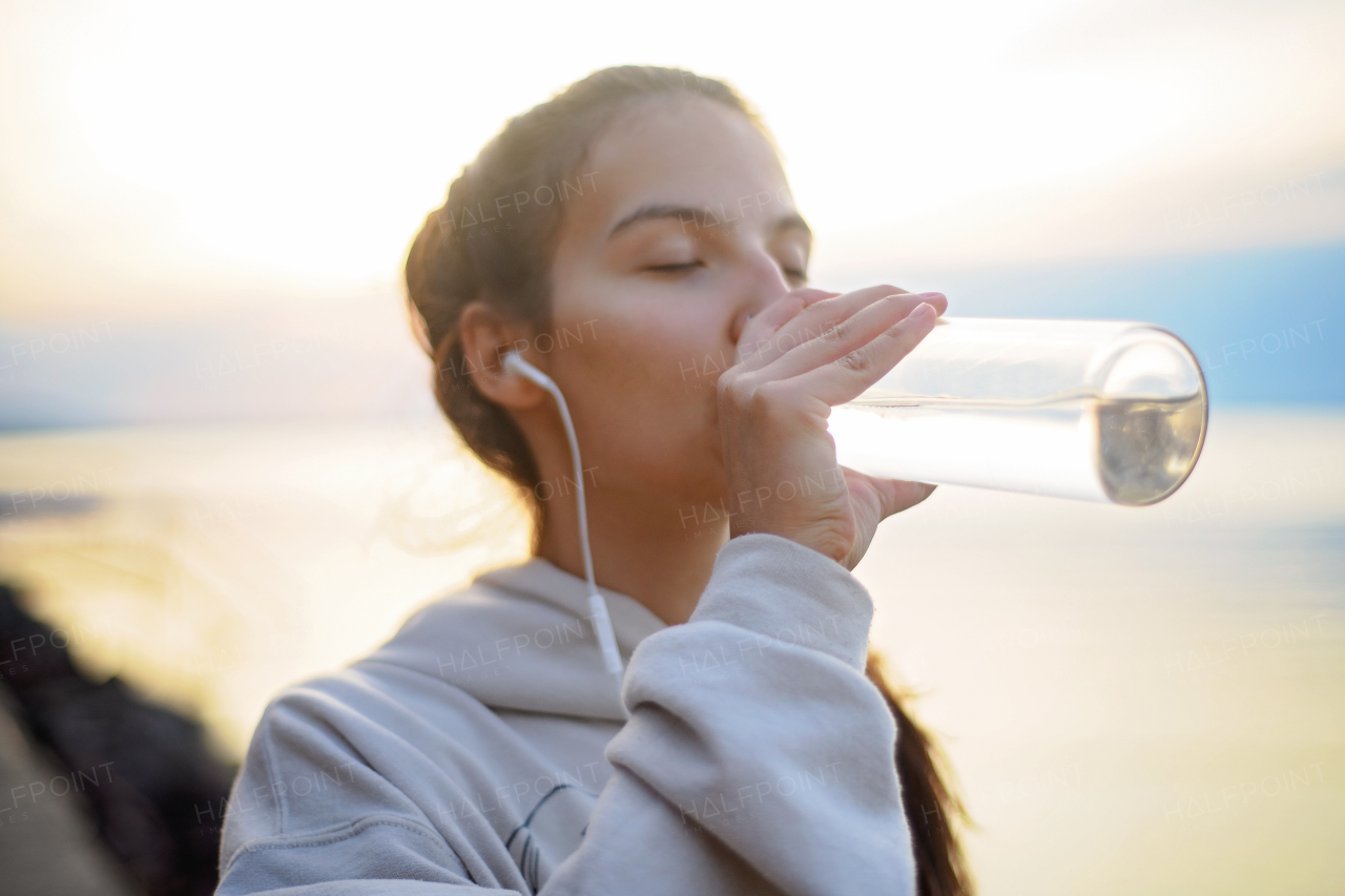 A young beautiful sportive girl listening to music and drinking water at sunrise by the lake.