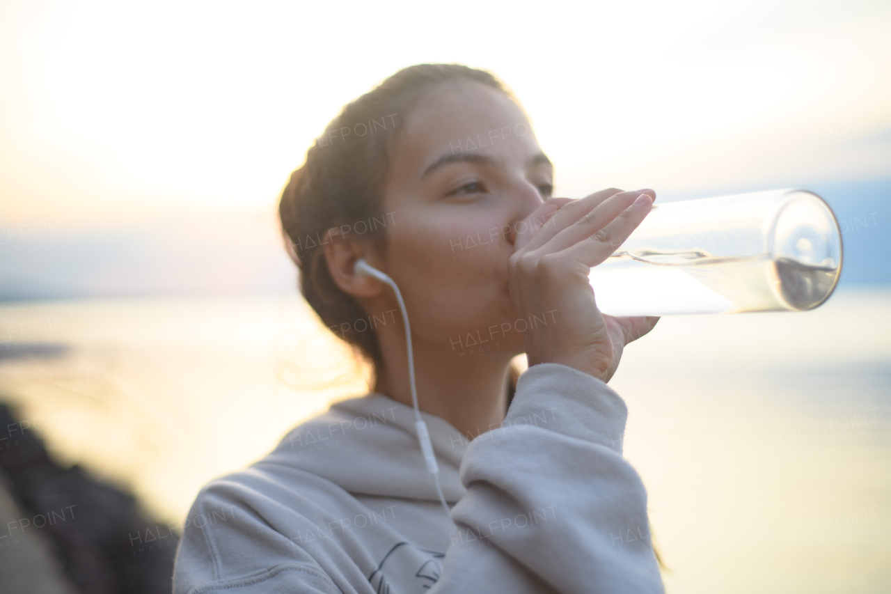 A young beautiful sportive girl listening to music and drinking water at sunrise by the lake.